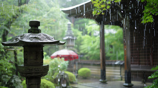 雨が降る和風の庭園と、手前に置かれた石灯籠、奥に見える木造の建物と赤い野点傘